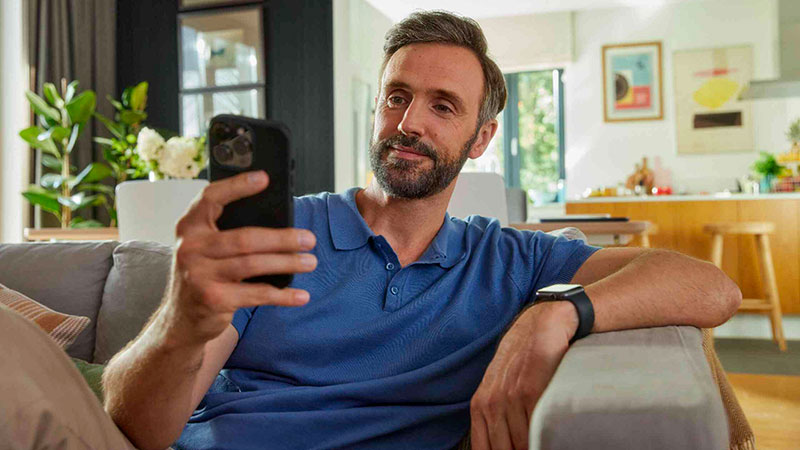 Vediamo un uomo seduto sul divano mentre guarda lo smartphone in ambiente domestico